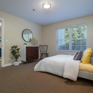 A cozy, minimalist bedroom with a neatly made bed, a chest of drawers with a round mirror above it, and a window letting in natural light.