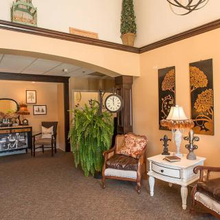 A cozy lobby area featuring comfortable chairs, artwork on the wall, decorative plants, and a table with a lamp and clock.