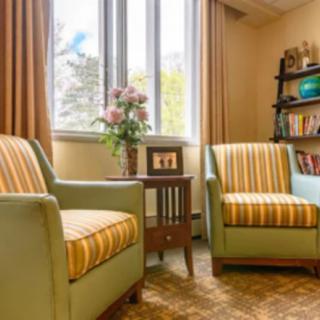 A cozy living room with two striped chairs, a small wooden table, flowers, and a bookshelf by the window.