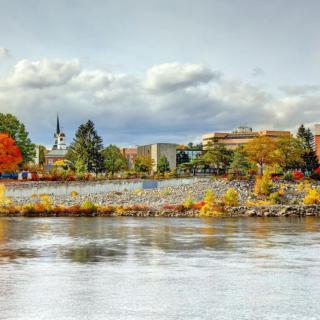A scenic view of a river lined with colorful autumn trees and buildings under a cloudy sky.