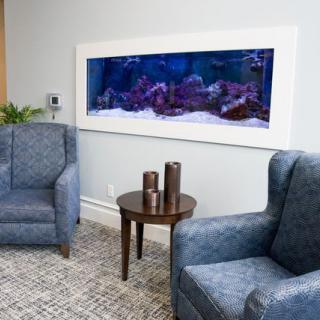 A cozy living room setting featuring two blue armchairs, a small round table with decorative items, and a large wall-mounted aquarium.
