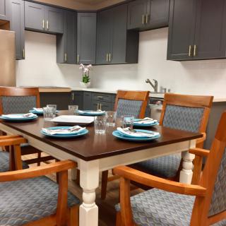 A modern dining room with a wooden table set for four, surrounded by matching chairs, and a stylish kitchen in the background.