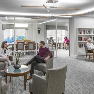 A modern office setting with people engaging in conversation and working in a comfortable seating area with glass walls and bookshelves.