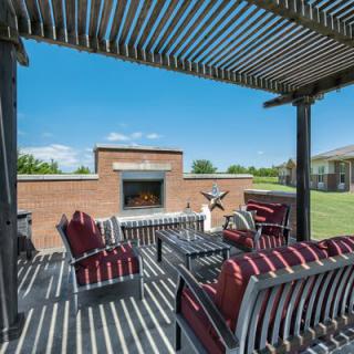 A sunny outdoor patio with a pergola, furnished with striped chairs and a central fireplace.