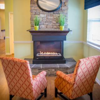 A cozy living room setup featuring two patterned chairs facing a stone fireplace, adorned with plants and a mirror.