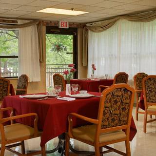 A well-arranged dining room with tables and chairs, featuring red tablecloths and large windows with curtains.