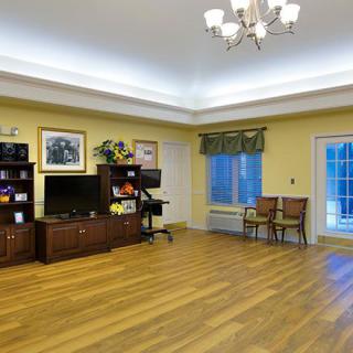 A spacious living room with wooden floors, a television on a shelf, and two chairs near a window.