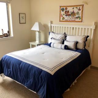 A cozy bedroom featuring a neatly made bed with blue and white bedding, a bedside lamp, artwork on the wall, and natural light from a window.