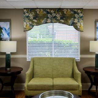 A cozy living room setting featuring a green couch, decorative lamps, and floral window treatments.