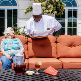 A chef serves a woman seated on an outdoor sofa with a drink and snack on the table.