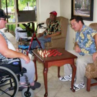 Three men engaged in a chess game indoors, one seated in a wheelchair.