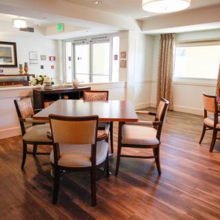 A cozy dining area with tables and chairs set on wooden flooring, illuminated by natural light filtering through large windows.