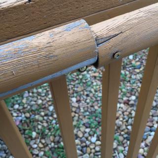A close-up of a cracked wooden railing with visible screws, set against a background of gravel.