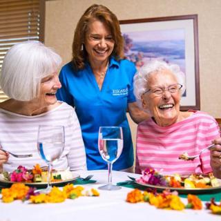 A caregiver sharing a joyful moment with two elderly women during a meal.