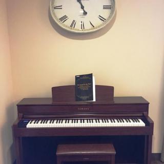 A piano with a music book placed on its stand, set under a wall clock.