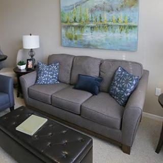 A cozy living room with a grey sofa adorned with patterned cushions, a decorative artwork on the wall, and a leather coffee table in front.