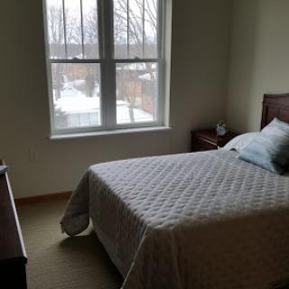 A cozy bedroom with a bed, nightstand, and large window letting in natural light.