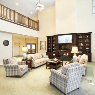 A spacious living room with a high ceiling, featuring comfortable sofas and armchairs arranged around a fireplace and shelves.