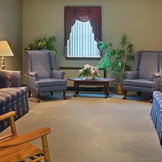 A cozy living room with blue armchairs, a sofa, and a central table adorned with flowers, surrounded by potted plants.