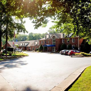 A sunlit brick building with a parking area surrounded by trees.