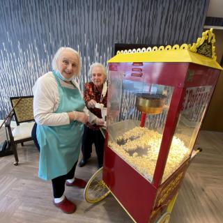 Two elderly women smiling next to a popcorn machine indoors.