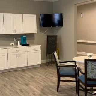 A modern kitchenette area with white cabinets, a small dining table and chairs, and a wall-mounted television.