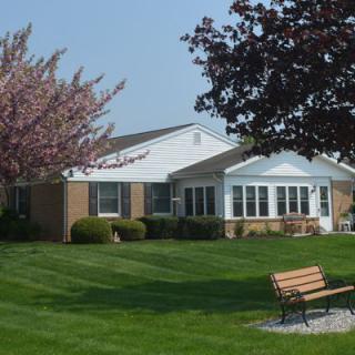 A suburban house with a well-maintained lawn, surrounded by trees and featuring a bench in the foreground.
