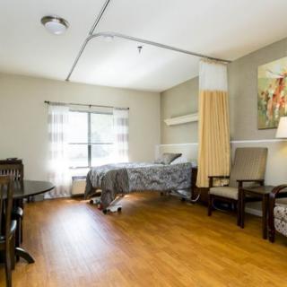 A cozy nursing home bedroom with a single bed, wood flooring, and ample natural light from a large window.