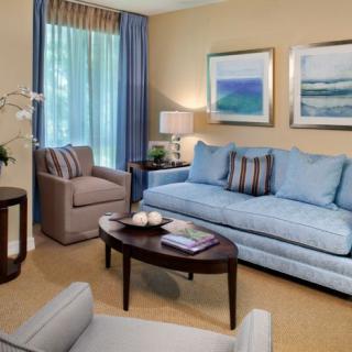 A cozy living room with a blue sofa, armchair, and a wooden coffee table, adorned with artwork and curtains.
