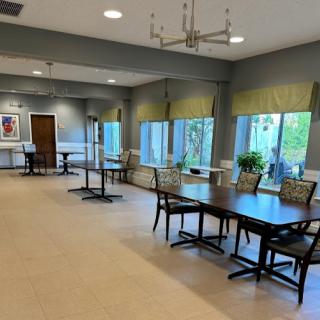 A spacious and well-lit dining area featuring neatly arranged tables and chairs, large windows with greenery outside.