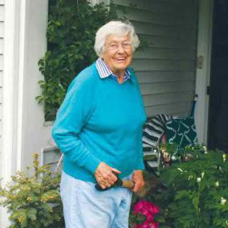 An elderly woman smiling while standing in a garden near a porch.