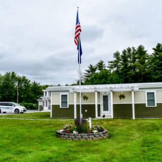 A small house with an American flag on a flagpole, surrounded by a well-maintained lawn and trees under a cloudy sky.