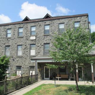 A three-story stone building with several windows, surrounded by greenery under a blue sky.