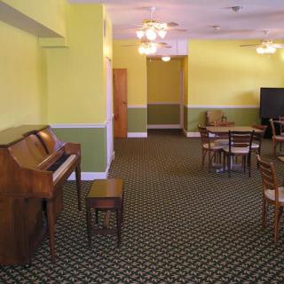 A room with a piano, tables, and chairs, featuring carpeted flooring and yellow walls.