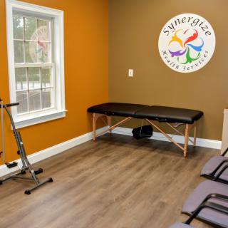 A therapy room with an exercise bike, massage table, and chairs, featuring a colorful logo on the wall.