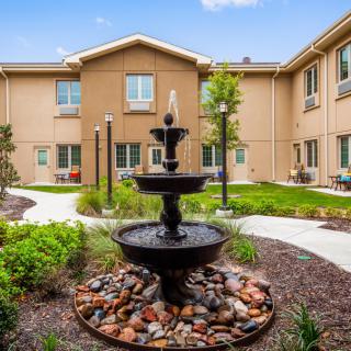 A serene courtyard featuring a decorative fountain surrounded by greenery and a multi-story building.