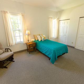 A cozy bedroom with a teal bedspread, large windows, and simple furnishings.