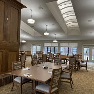A spacious dining room with neatly arranged tables and chairs, illuminated by ceiling lights and natural light from large windows.