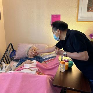 A visitor wearing a mask comforts an elderly patient in a hospital bed.