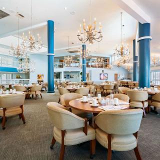 A spacious and elegantly designed restaurant with chandeliers hanging from the ceiling, set up with round tables and plush chairs.