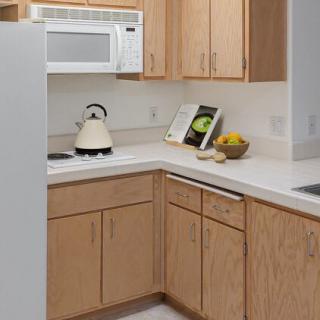 A modern kitchen with wooden cabinets, a white countertop, and various kitchen items including a microwave, kettle, and fruit bowl.
