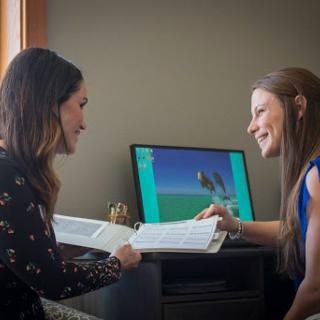 Two women engaging in a discussion while reviewing materials beside a computer.