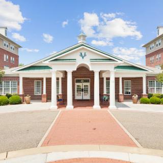 A welcoming entrance to a modern building, featuring classical columns and landscaped surroundings under a bright blue sky.