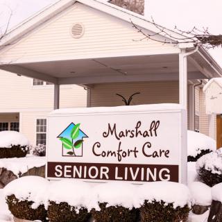 A snow-covered sign for Marshall Comfort Care Senior Living in front of a building.