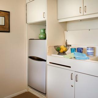 A compact kitchen corner with white cabinets, a small refrigerator, and a tidy countertop featuring a fruit bowl and some kitchen items.