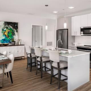 A modern kitchen and dining area with sleek white cabinetry, stainless steel appliances, and an island with bar stools.