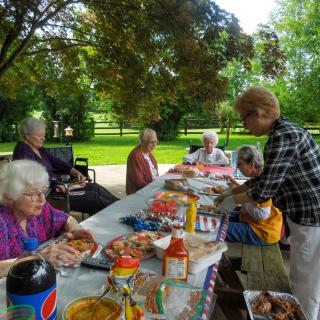 A group of people enjoying a picnic at a park table, surrounded by trees and an assortment of food and drinks.