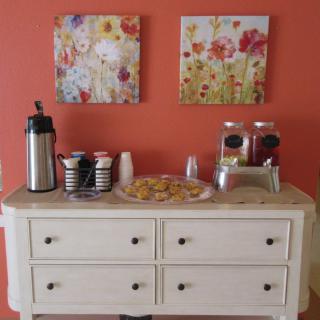 A cozy refreshment station with coffee, cups, and pastries displayed on a white buffet table against a warm orange wall adorned with floral artwork.