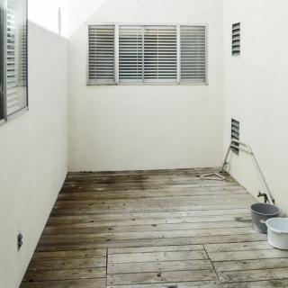 A minimalist outdoor patio with wooden flooring and white walls, featuring window shutters.