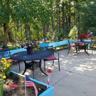 A charming patio area with tables and chairs surrounded by vibrant flower planters and small flags, set against a backdrop of lush trees.